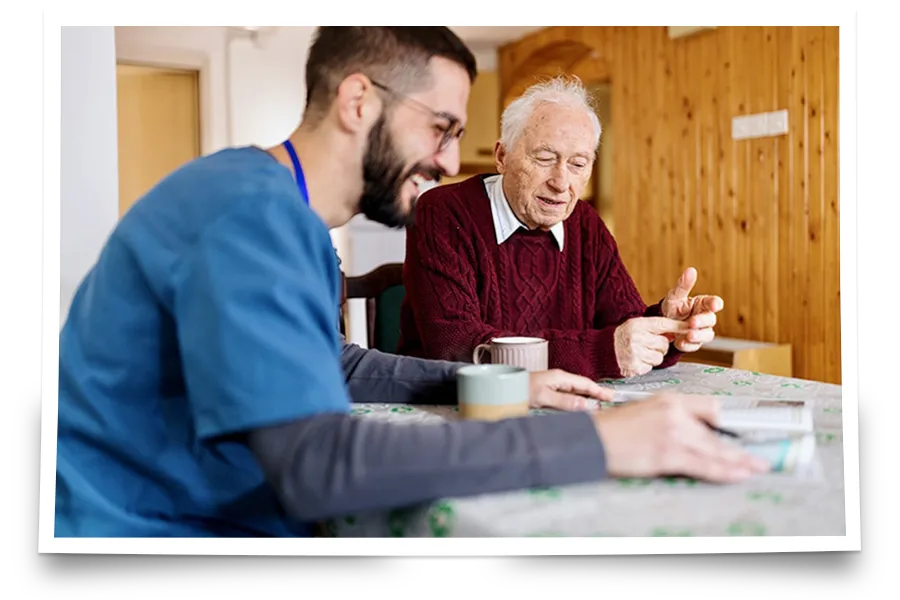 A young male caregiver in blue scrubs sits at a table with an elderly man in a red sweater; both are smiling and talking over drinks and a pack of cards.