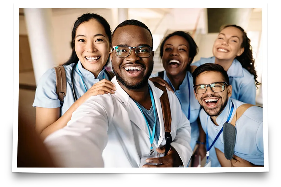 Five young adults in medical scrubs and badges smile and pose together for a group selfie indoors.