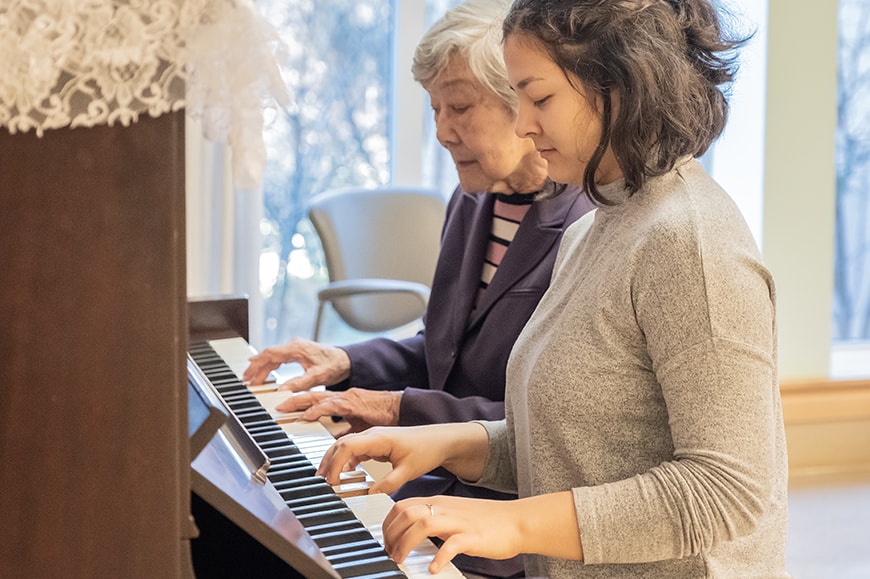 An older woman and a younger woman play a piano together in a bright room, both focused on the keys.