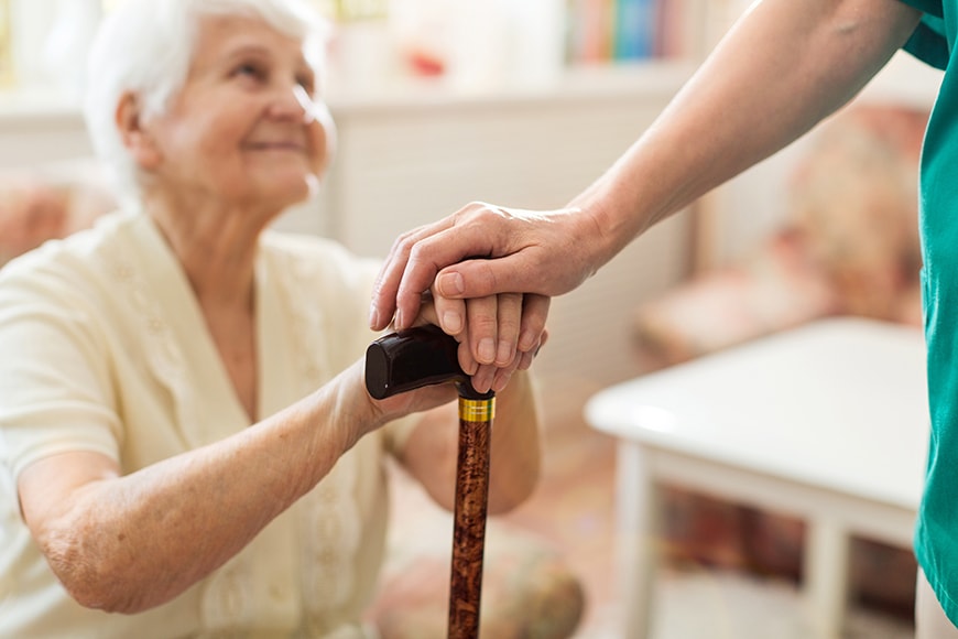 An elderly woman holding a cane receives support from a standing person, whose hand rests gently on hers.