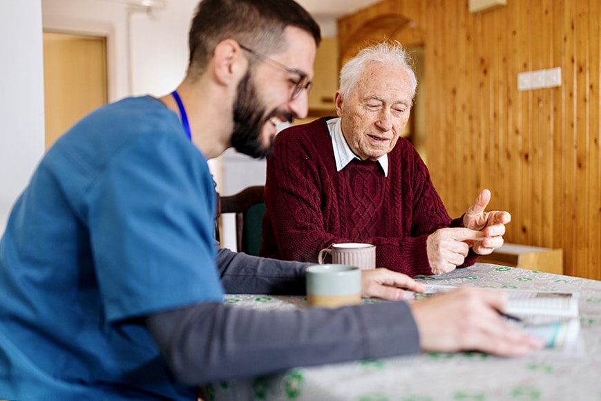 A healthcare worker and an elderly man sit at a table, talking and smiling, with mugs in front of them in a cozy indoor setting.
