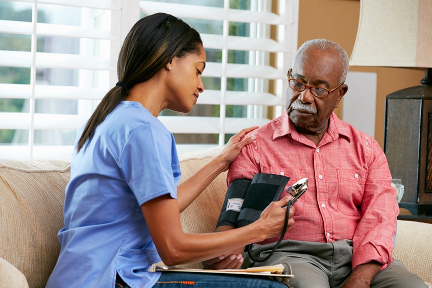 A healthcare worker measures an older man's blood pressure at home. The man sits on a couch while the worker checks the monitor and holds his arm.