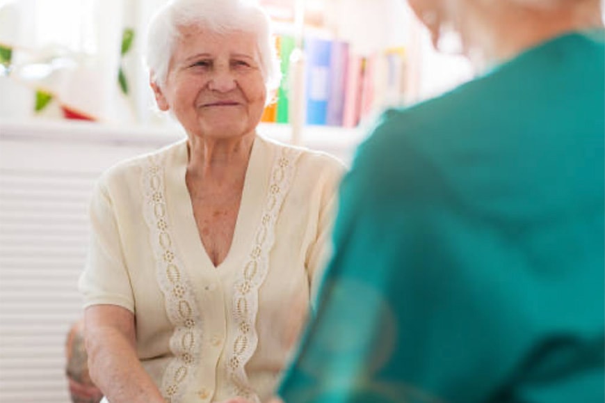 An elderly woman with short white hair smiles while sitting indoors, facing another person in a teal outfit.
