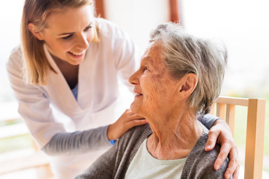 A young caregiver in uniform smiles and places a hand on the shoulder of an older woman sitting in a chair.