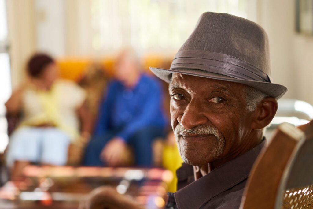 An older man wearing a gray hat sits in the foreground, looking at the camera, with two people talking on a couch in the blurred background.