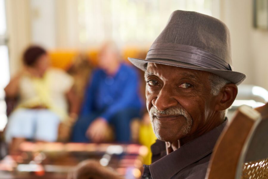 An older man wearing a gray hat sits in the foreground, looking at the camera, with two people talking on a couch in the blurred background.