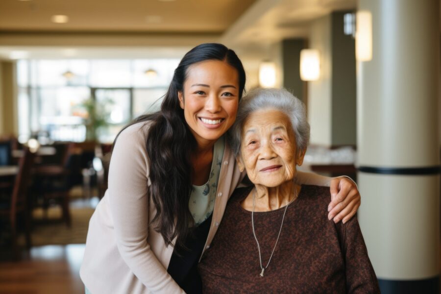 A younger woman with long hair smiles and embraces an older woman with short gray hair, both posing together indoors in a well-lit room.
