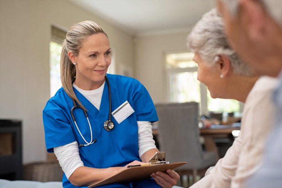A healthcare professional in blue scrubs holds a clipboard and talks with two older adults in a home setting.