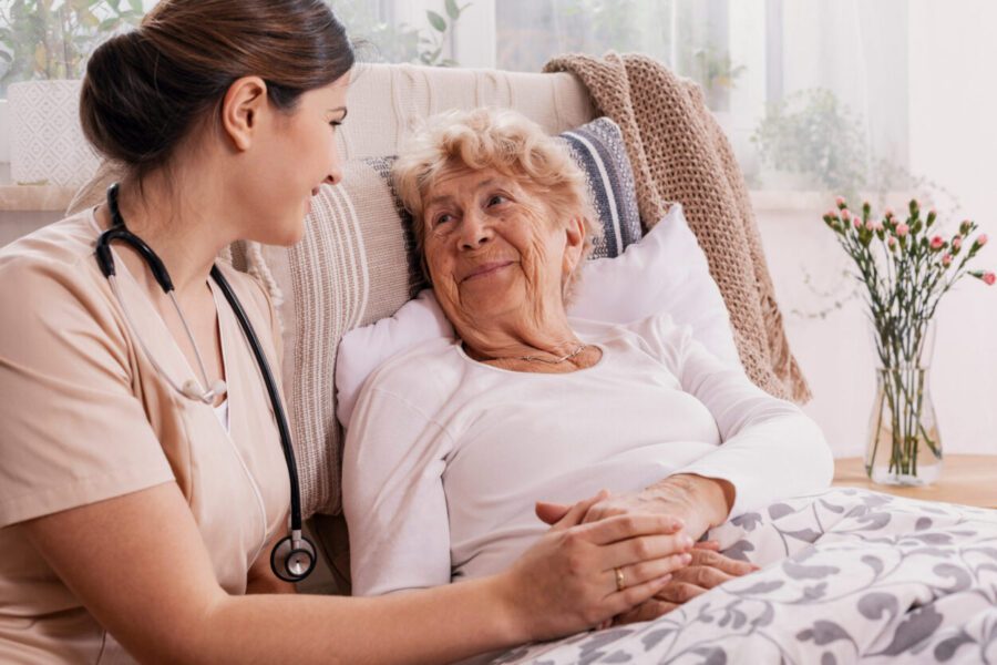 A nurse with a stethoscope holds hands with an elderly woman lying in bed, both smiling at each other in a cozy, well-lit room.