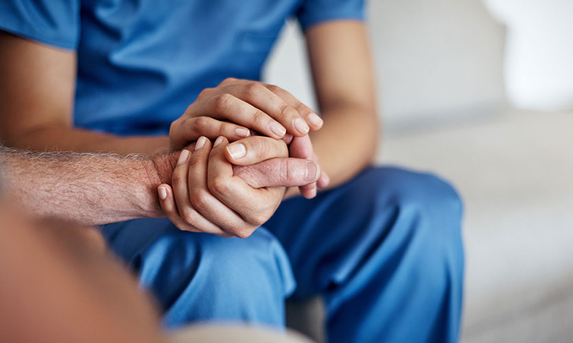 A person in blue scrubs sits and gently holds the hand of another person, offering support and comfort.