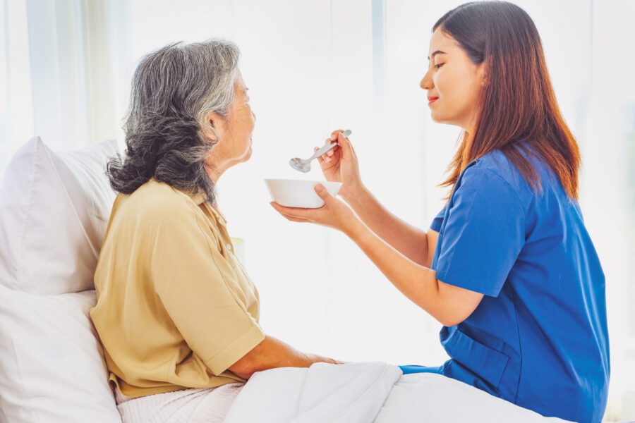 A nurse in blue scrubs feeds an elderly woman sitting up in bed with a spoon and bowl in a brightly lit room.