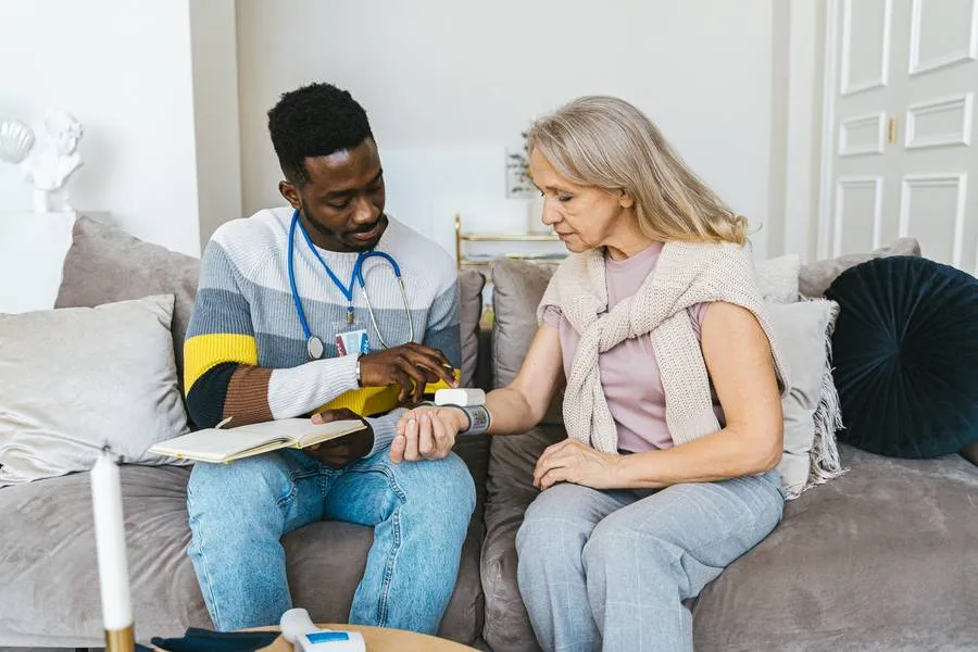 A healthcare professional checks the blood pressure of an older woman sitting on a couch during a home visit.